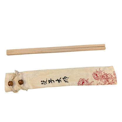 A pair of wooden chopsticks placed above their matching beige fabric sleeve, which features black writing and red floral illustrations, on a plain white background.