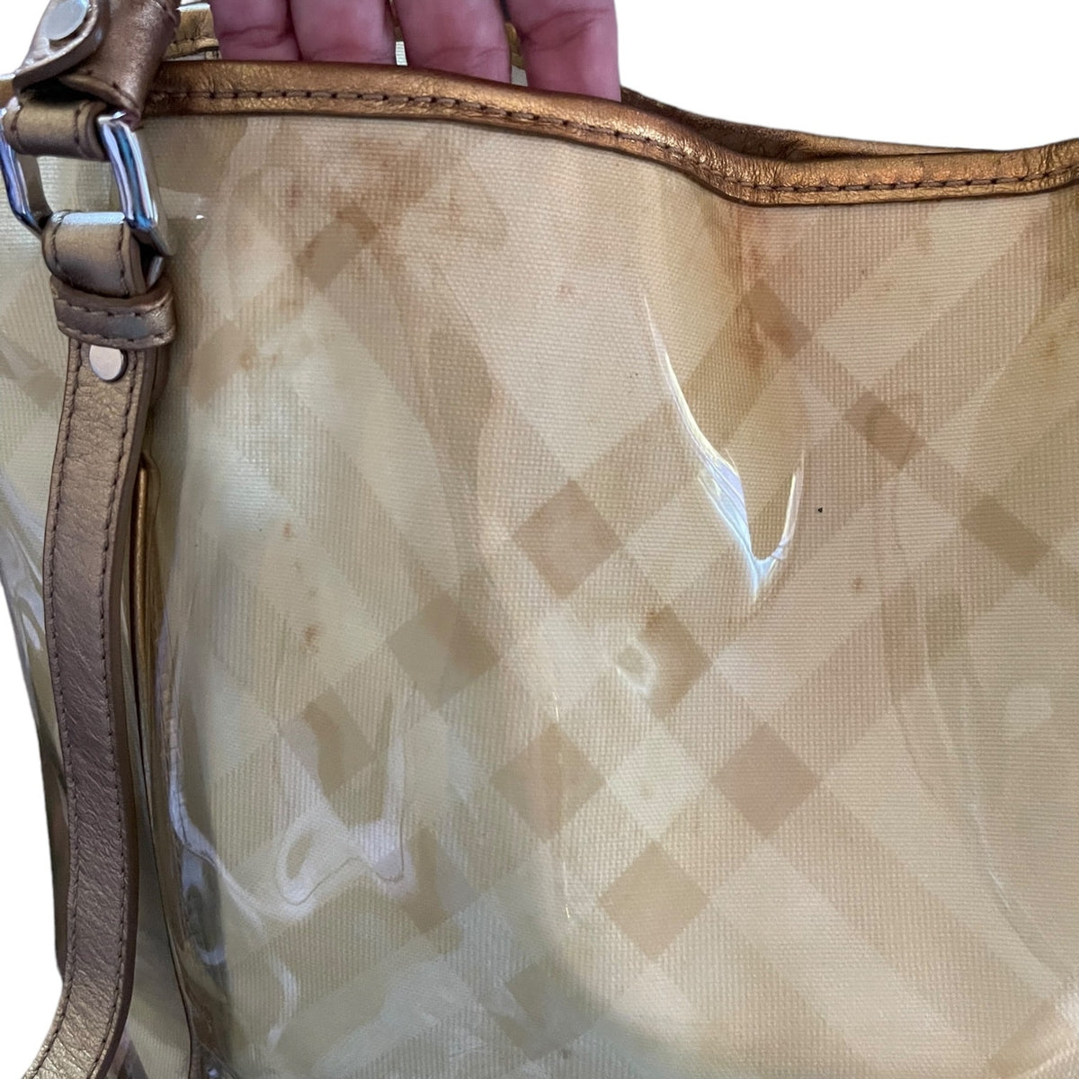 A close-up of a translucent beige and tan checkered handbag with a gold leather strap and trim, held by a hand at the top.