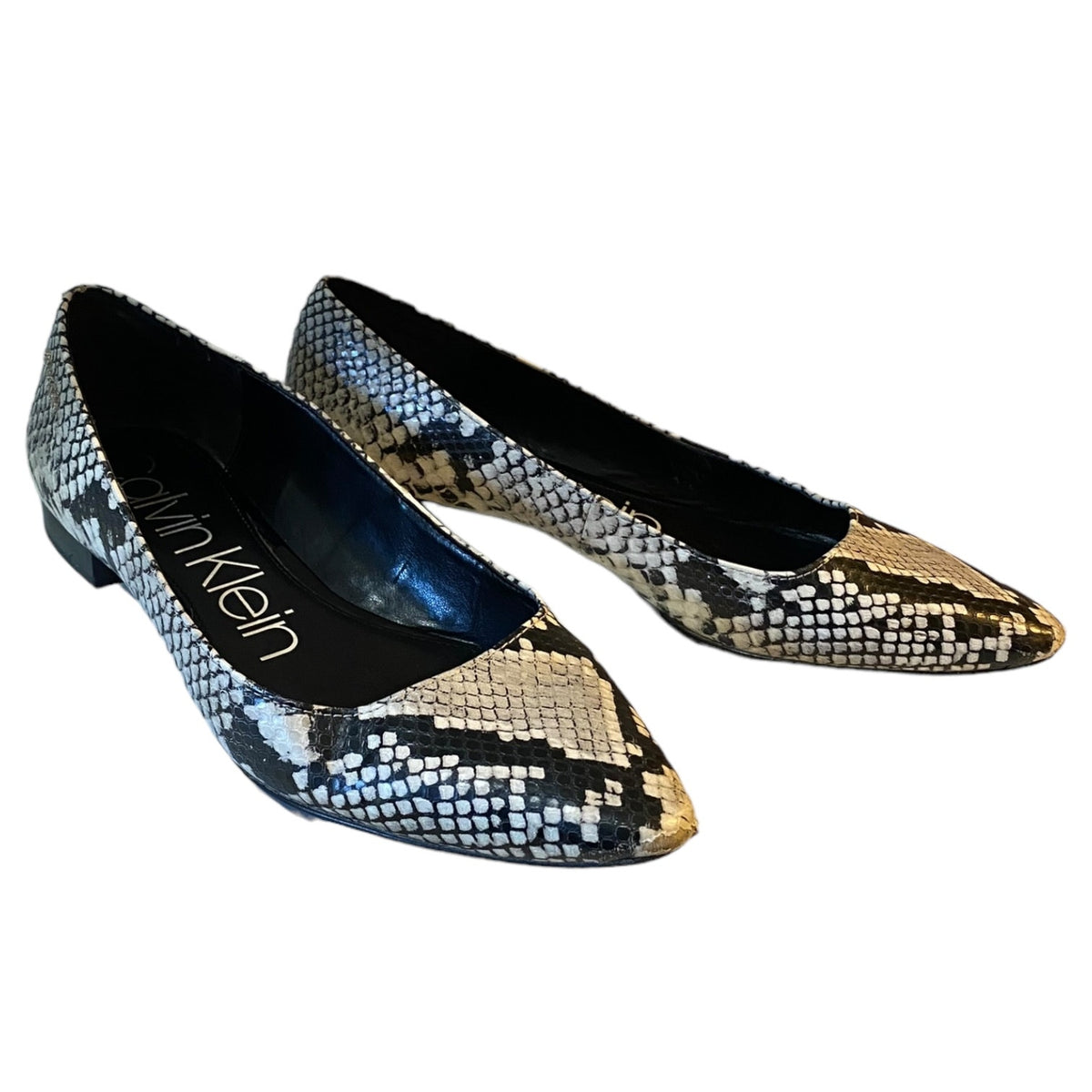 A pair of Calvin Klein pointed-toe flat shoes featuring a black, gray, and beige snakeskin pattern, shown on a plain white background.