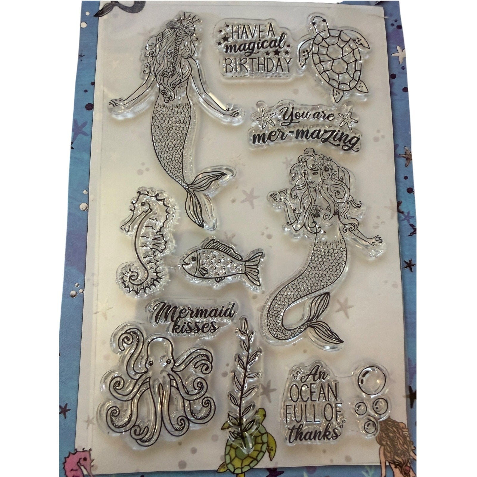 A sheet of clear acrylic or rubber stamps featuring mermaid, seahorse, fish, octopus, turtle, bubble, and decorative text designs, displayed on a patterned blue-and-white backing card.