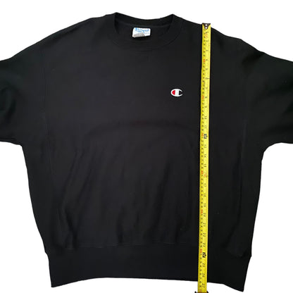 Black sweatshirt with a small logo on a white background, measured by a yellow tape measure.