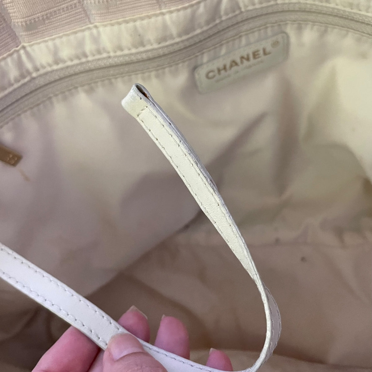 Close-up of a Chanel handbag interior with a cream-colored leather strap held by a hand, showing the CHANEL label stitched inside the lining.