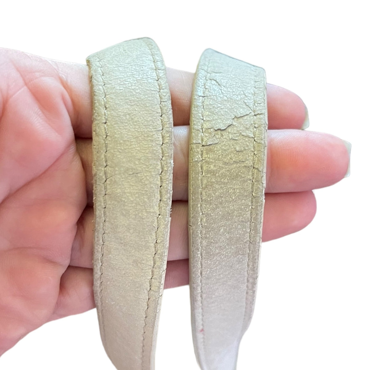 A close-up of a hand holding two off white beige leather straps side by side against a plain white background, showing the texture and condition of the straps.