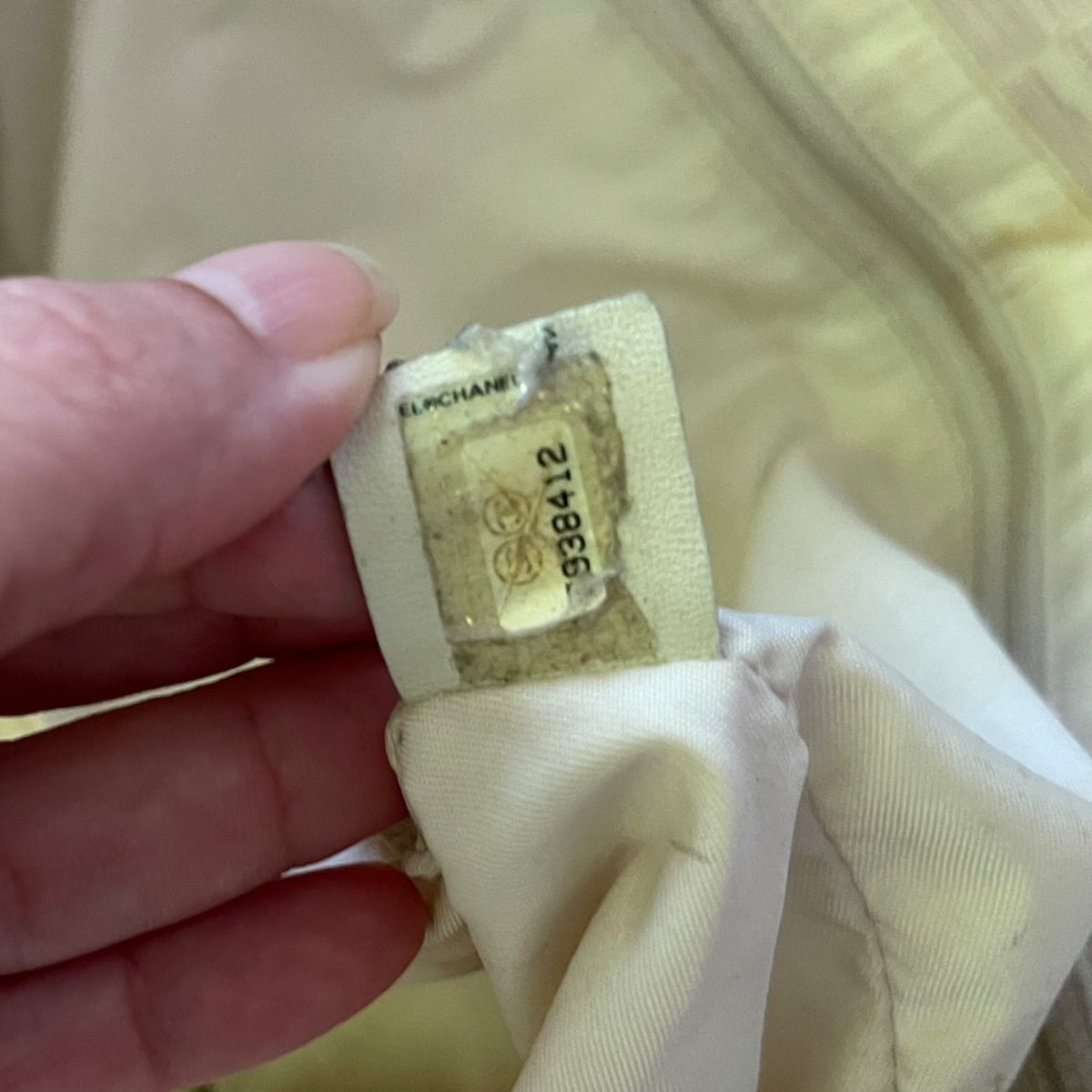 A close-up of a hand holding a beige fabric tag with a clear plastic window showing the printed Chanel logo and item number .