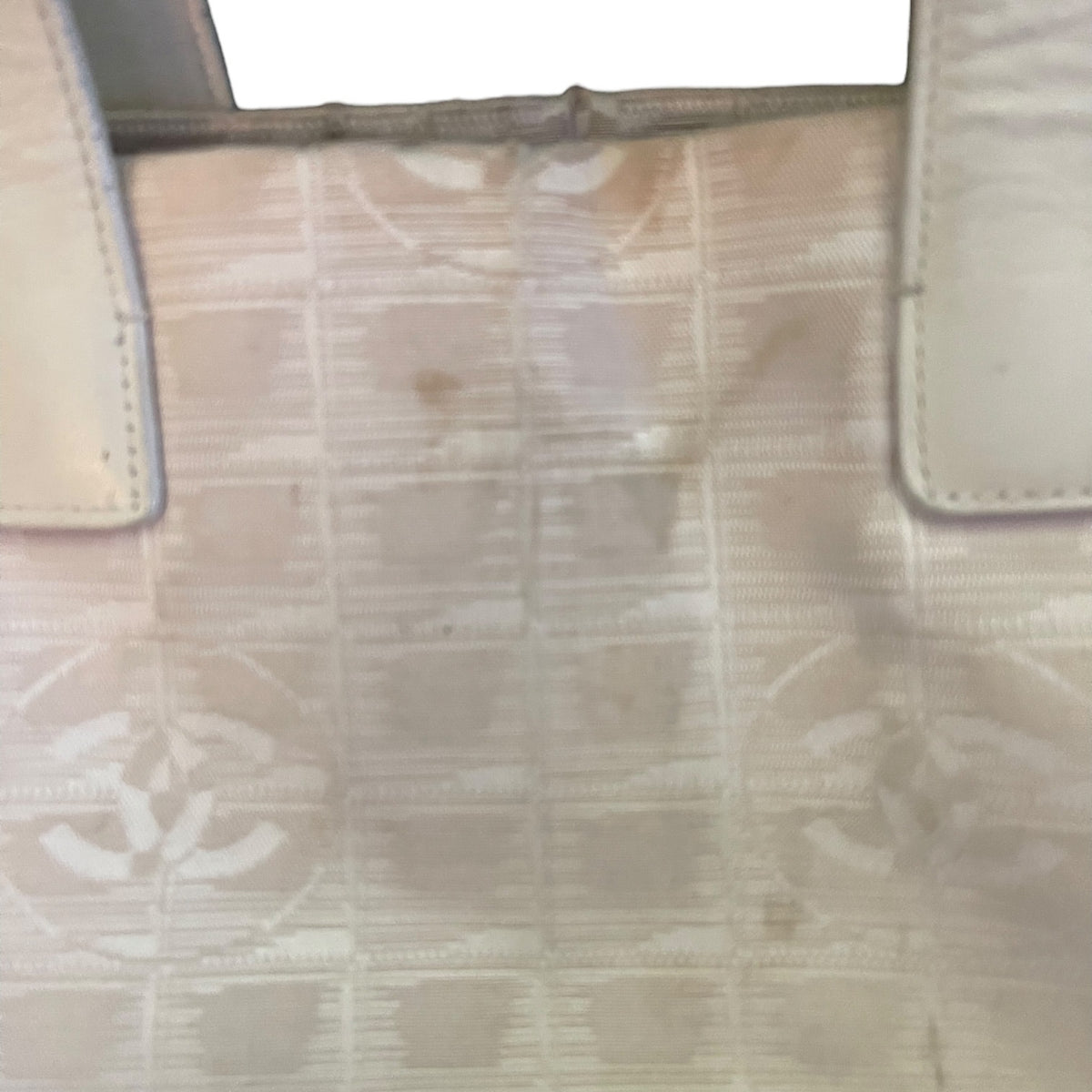A close-up of a beige and off-white patterned handbag surface with visible scuffs, stains, and wear, showing part of the strap attachment and a faint repeating monogram-like pattern.