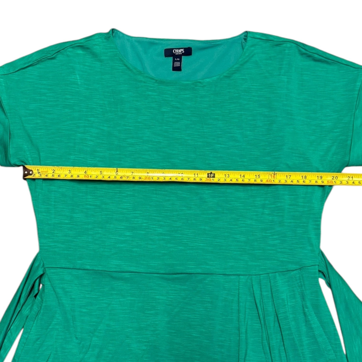 A green short-sleeve dress laid flat on a white background with a yellow measuring tape stretched horizontally across the chest to show the width.