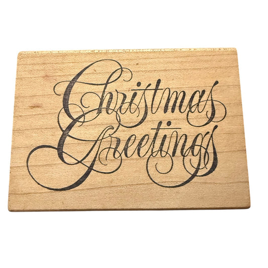 A rectangular wooden stamp with the words 'Christmas Greetings' written in large black cursive script, shown on a plain white background.