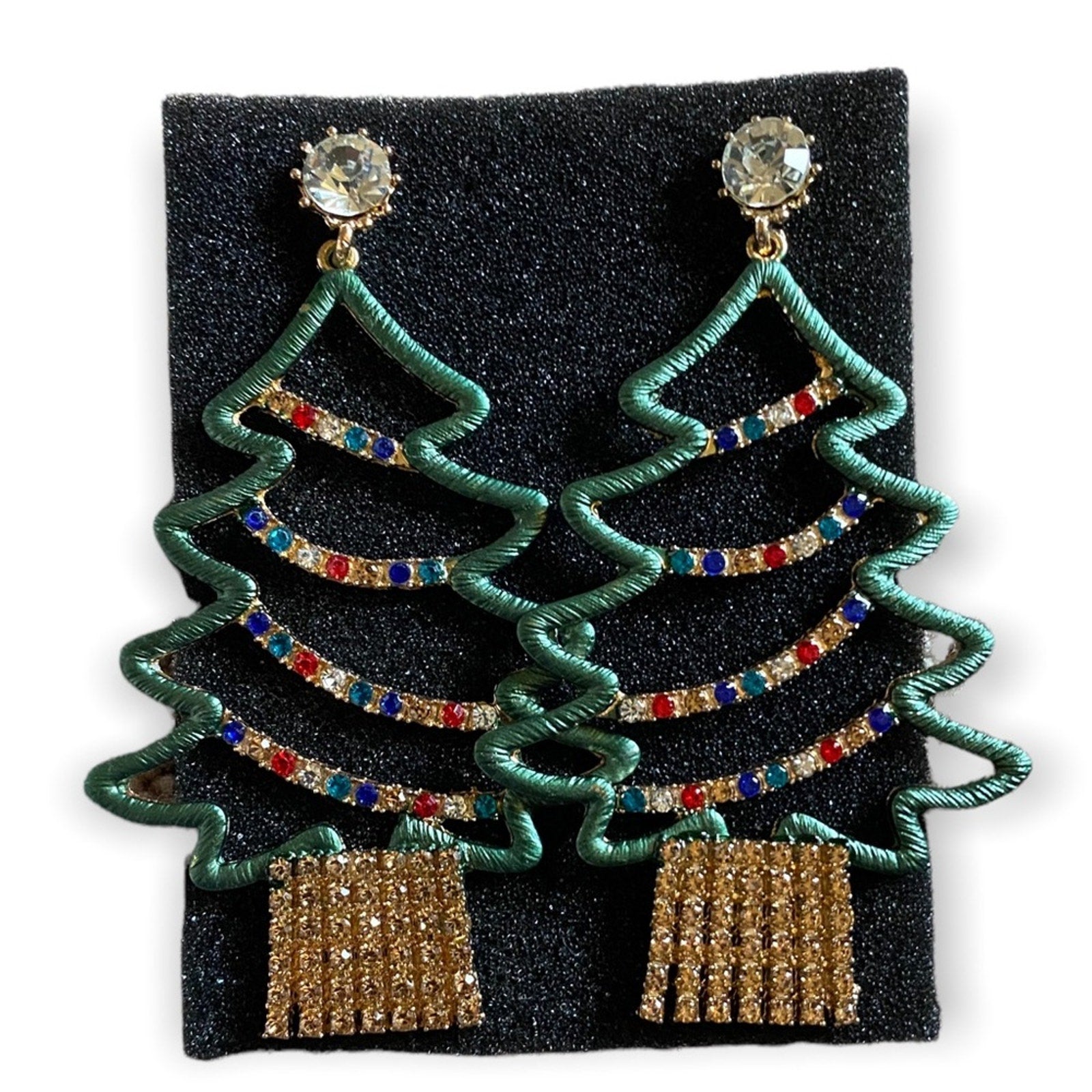 A pair of Christmas tree-shaped drop earrings with green wire outlines, multicolor rhinestone garlands, clear crystal studs, and gold dangling fringe, displayed on a black textured backing card against a white background.