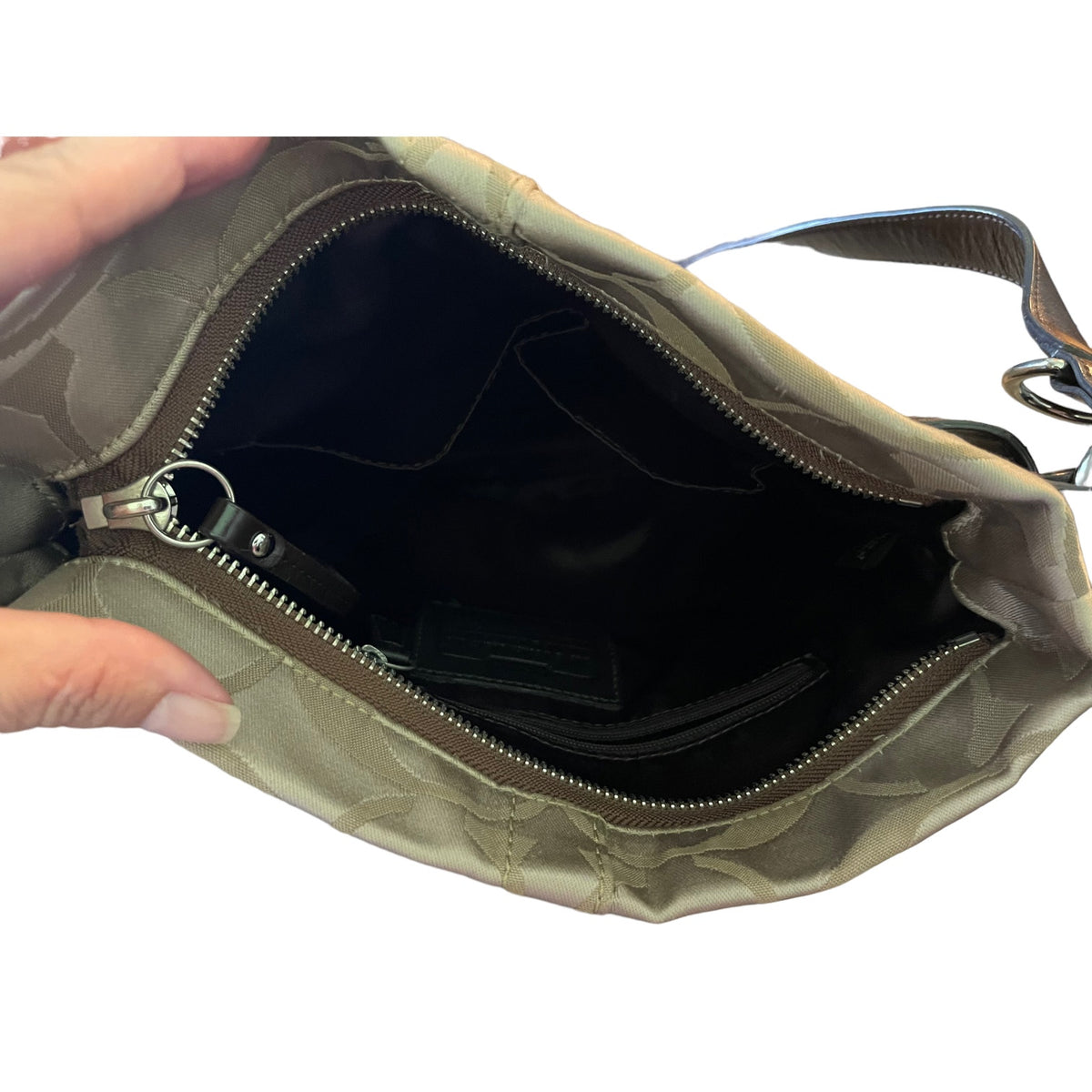 A close-up of a hand holding open a brown and beige patterned shoulder bag, showing the dark interior lining and zipper.
