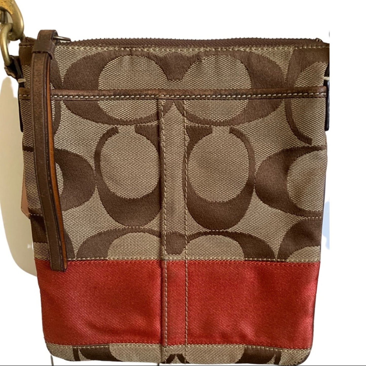 A Coach crossbody bag featuring the classic brown monogram canvas pattern, a brown leather wristlet strap, and a prominent orange horizontal stripe across the lower half.