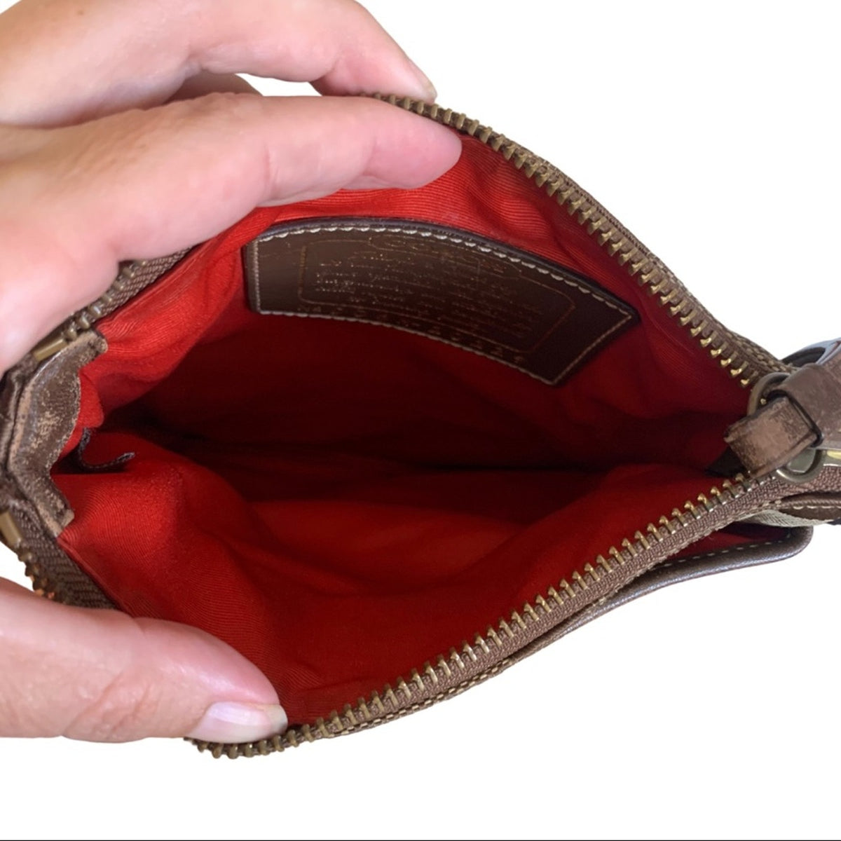 A close-up of a brown leather zippered pouch being held open by hands, showing a bright orange interior lining and a brown interior label.