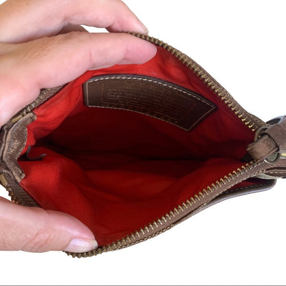A close-up of a brown leather zippered pouch being held open by hands, showing a bright orange interior lining and a brown interior label.