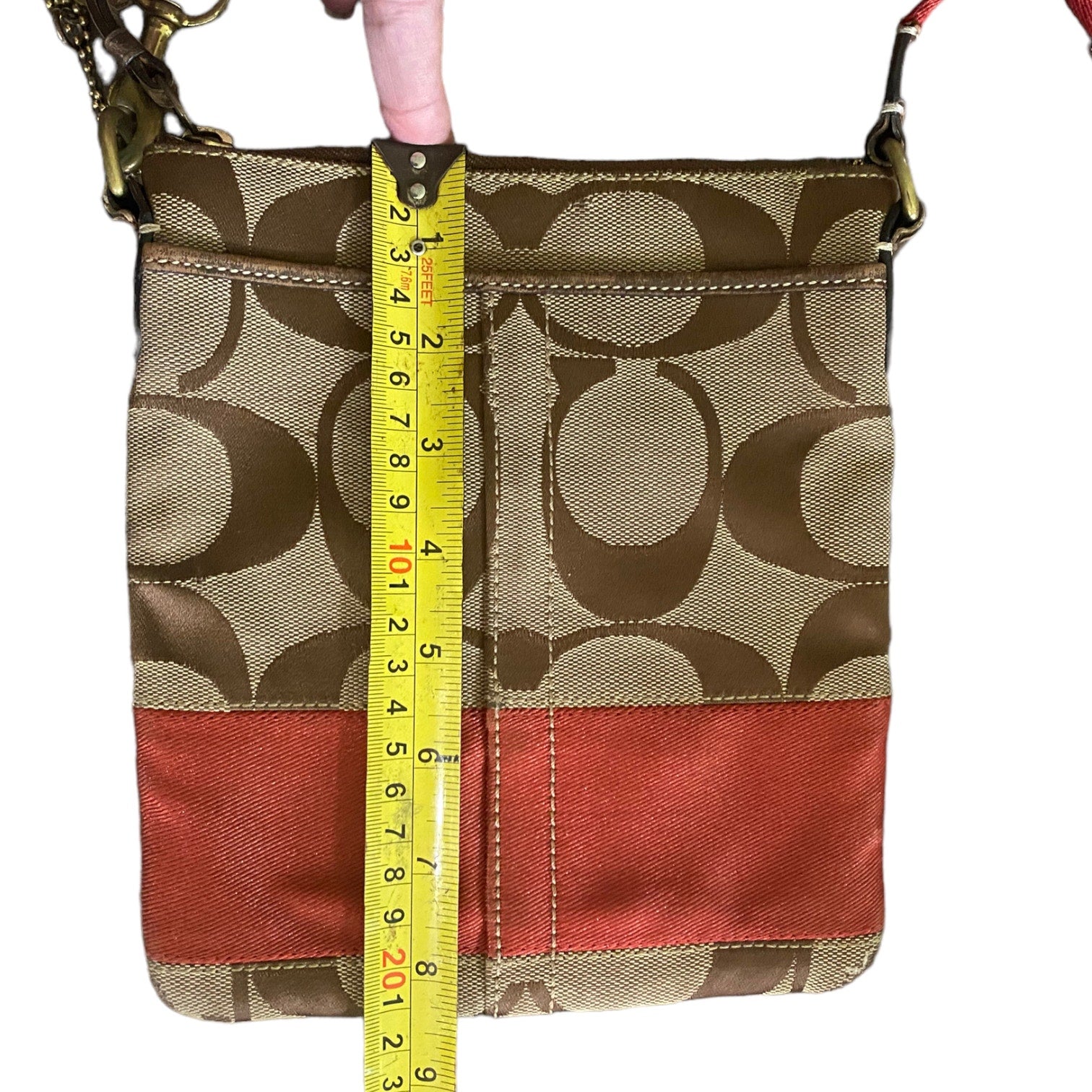 A Coach crossbody bag with the classic monogram pattern, featuring a yellow tape measure held vertically against it for size reference, on a white background.