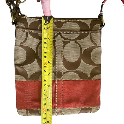 A Coach crossbody bag with the classic monogram pattern, featuring a yellow tape measure held vertically against it for size reference, on a white background.