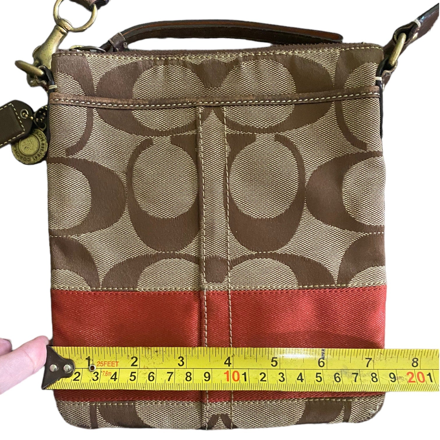 A Coach crossbody bag in the signature monogram canvas  shown with a yellow tape measure across the front to indicate size. A hand is holding the tape measure on the left.
