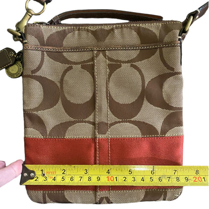 A Coach crossbody bag in the signature monogram canvas  shown with a yellow tape measure across the front to indicate size. A hand is holding the tape measure on the left.