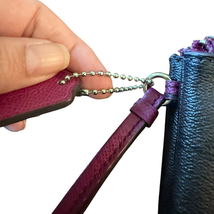 A close-up of a hand attaching a ball chain strap to a pink leather wristlet or pouch next to a black textured bag on a white background.
