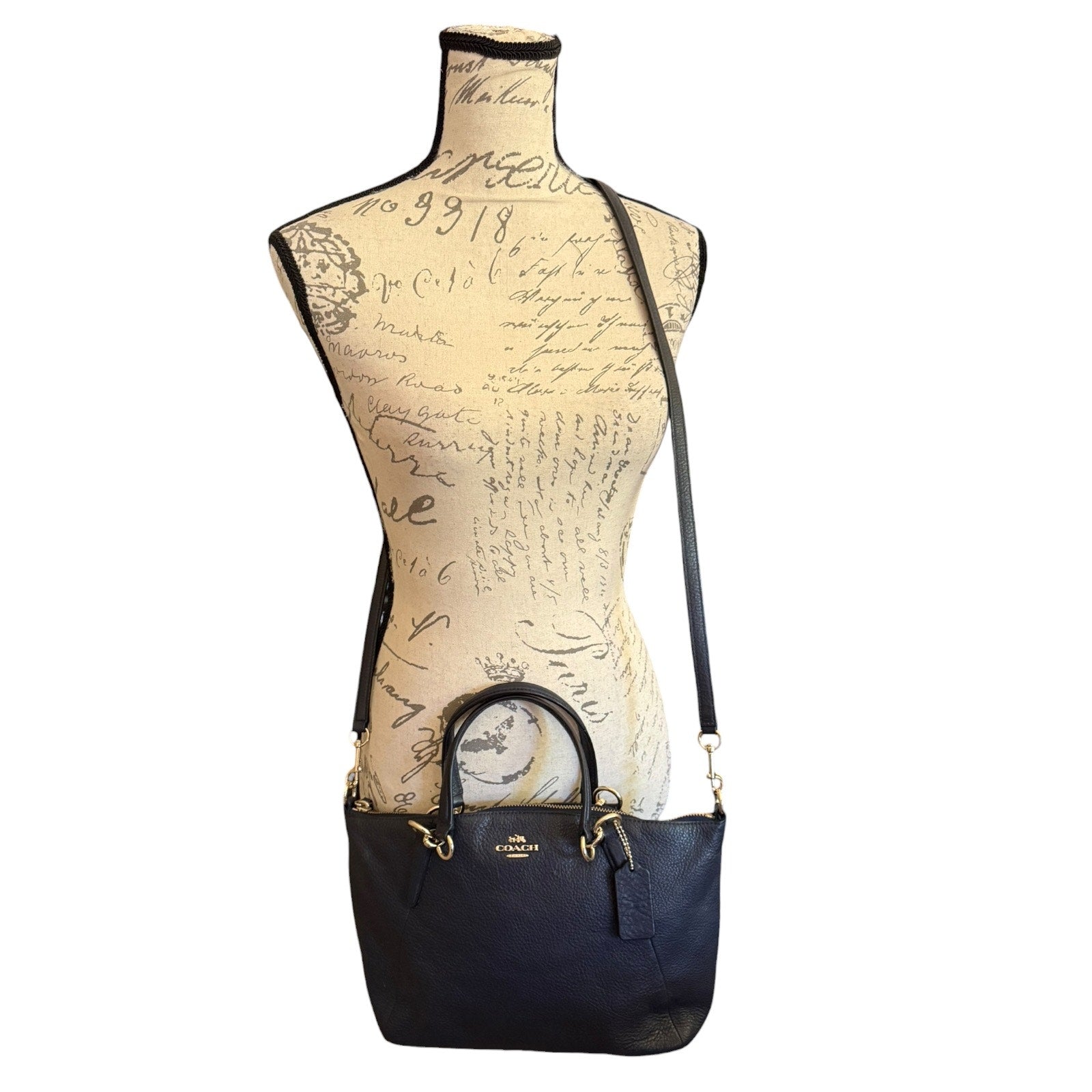A Coach handbag in black leather , displayed on a beige script-patterned dress form mannequin against a white background.