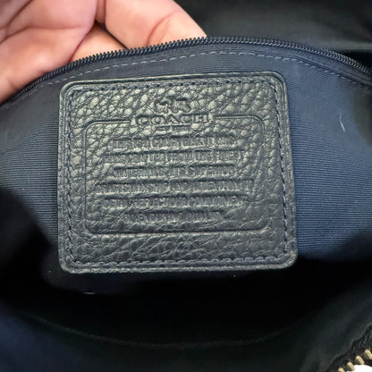 Close-up of a Coach bag interior showing a black pebbled leather brand patch with embossed lettering, held by a hand near the zipper.