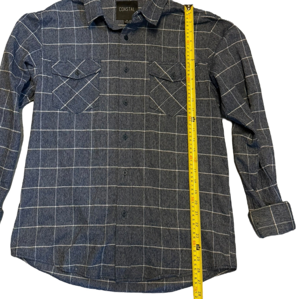 A blue plaid button-up shirt with a yellow measuring tape running vertically along the side to show the garment's length. 