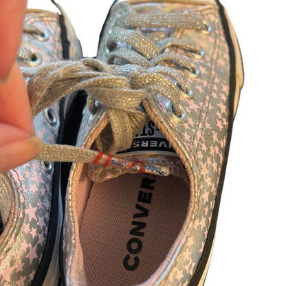Close-up of a Converse sneaker with a metallic silver and pink star pattern, featuring metallic laces and a visible hand adjusting one of the laces.