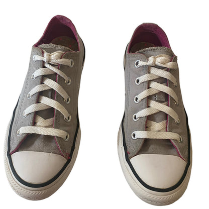 A pair of low-top canvas sneakers in a  gray color, featuring white laces, white rubber toe caps, and black rubber trim around the soles. The interior lining is a dark magenta.