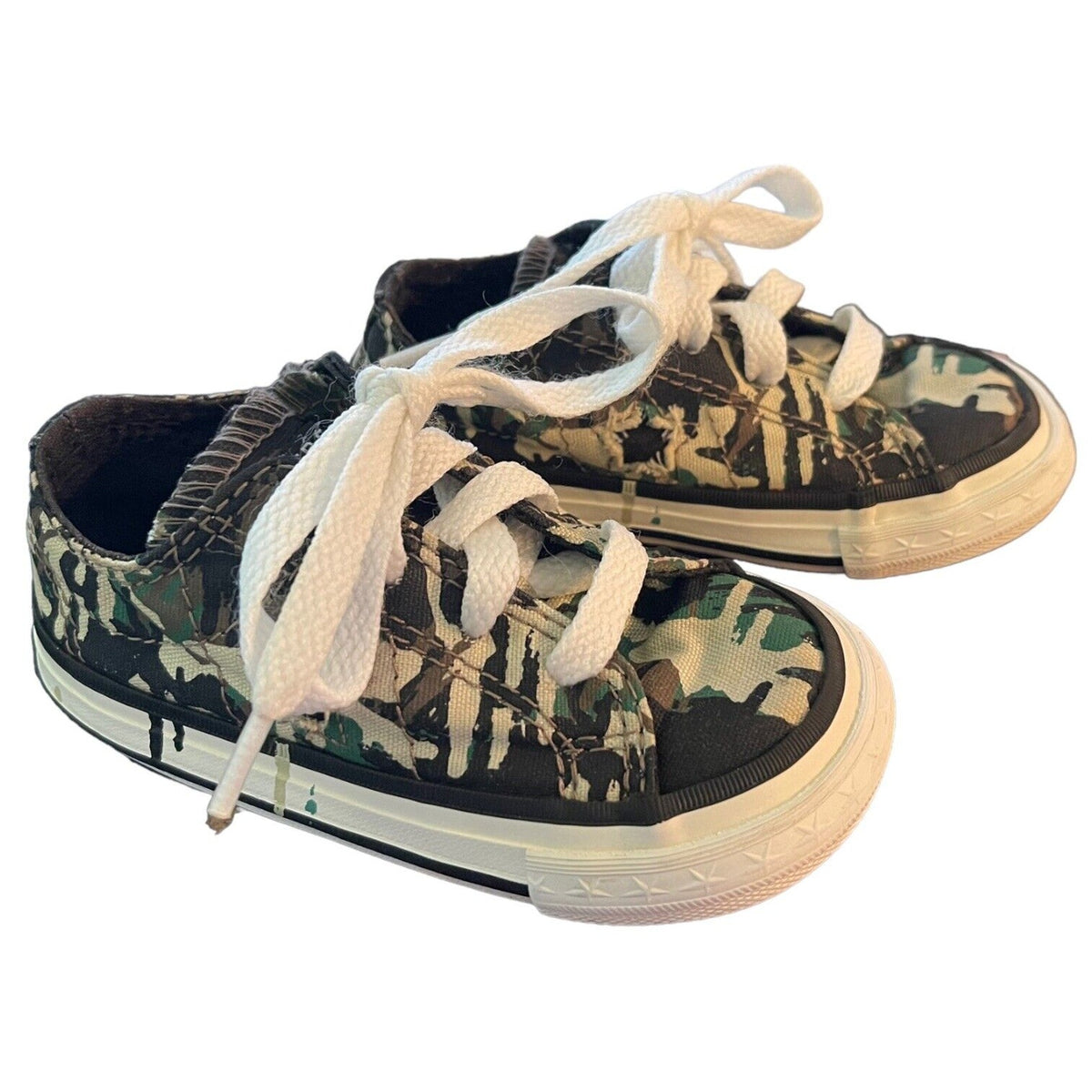A pair of low-top canvas sneakers with a green, black, and beige camouflage pattern, featuring white laces and off-white rubber soles, shown on a plain white background.