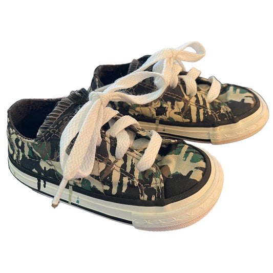 A pair of low-top canvas sneakers with a green, black, and beige camouflage pattern, featuring white laces and off-white rubber soles, shown on a plain white background.