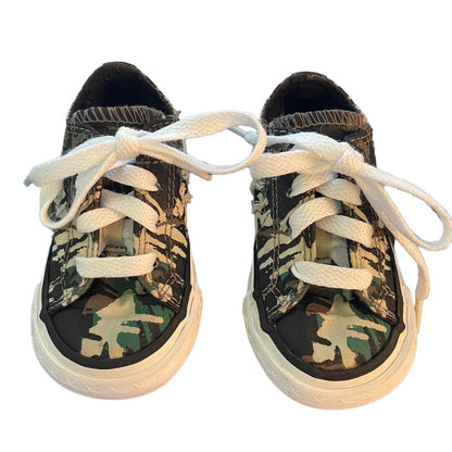 A pair of low-top canvas sneakers with a green, brown, and black camouflage pattern, featuring white laces and white rubber soles, shown against a plain white background.