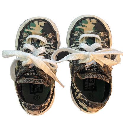 A pair of toddler Converse All Star sneakers with a camouflage pattern, featuring white laces tied in bows and white rubber soles, shown from a top-down view on a white background.