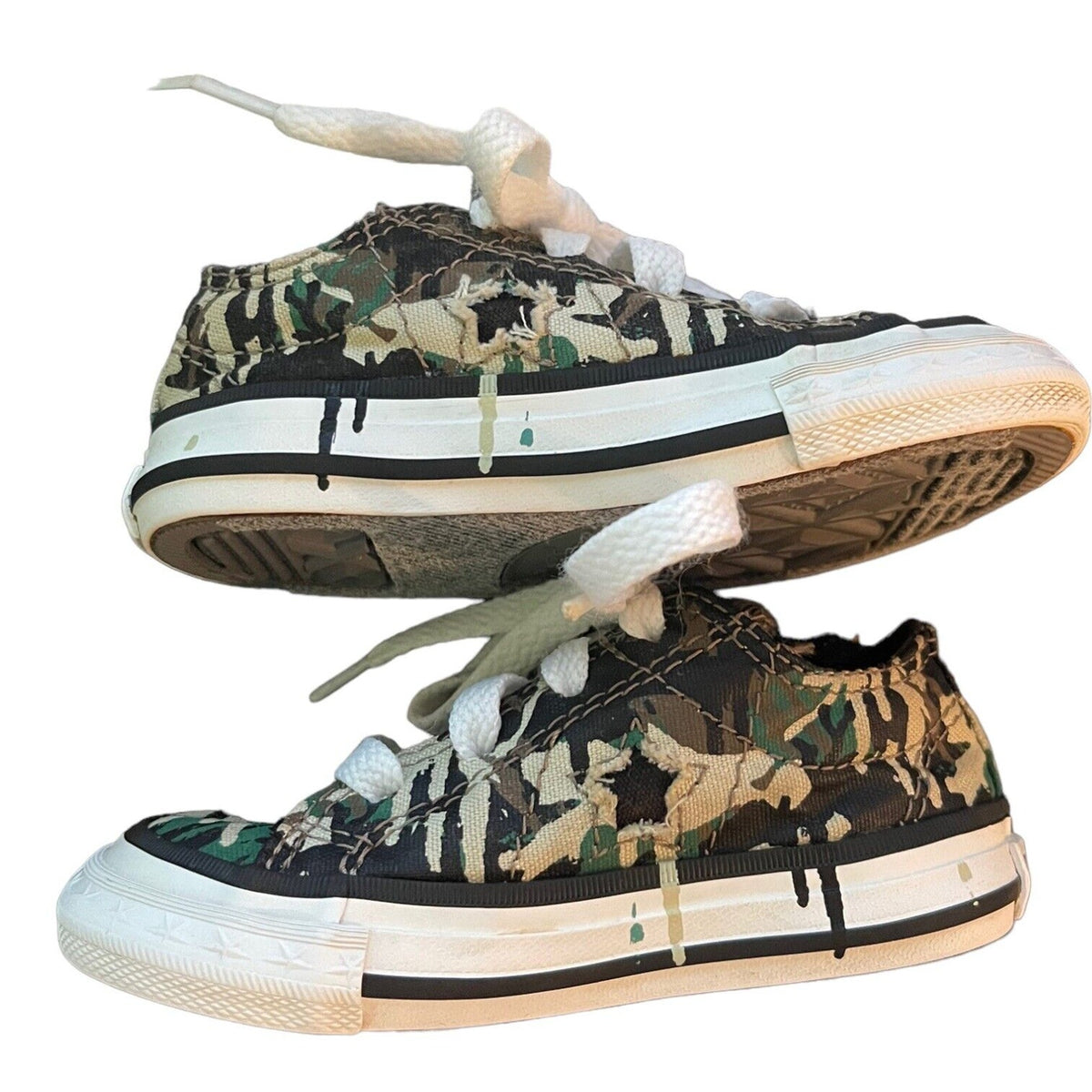 A pair of low-top canvas sneakers with a distressed camouflage pattern in green, brown, and beige, featuring white laces and white rubber soles with black detailing.