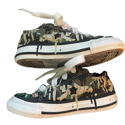 A pair of low-top canvas sneakers with a distressed camouflage pattern in green, brown, and beige, featuring white laces and white rubber soles with black detailing.