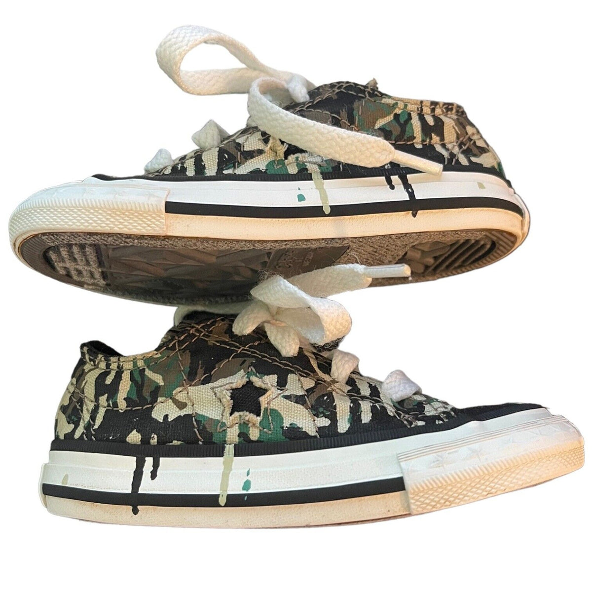 A pair of low-top canvas sneakers with a green, black, and beige camouflage pattern, featuring white laces and white rubber midsoles with black stripes, shown against a plain white background.