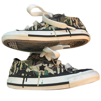A pair of low-top canvas sneakers with a green, black, and beige camouflage pattern, featuring white laces and white rubber midsoles with black stripes, shown against a plain white background.