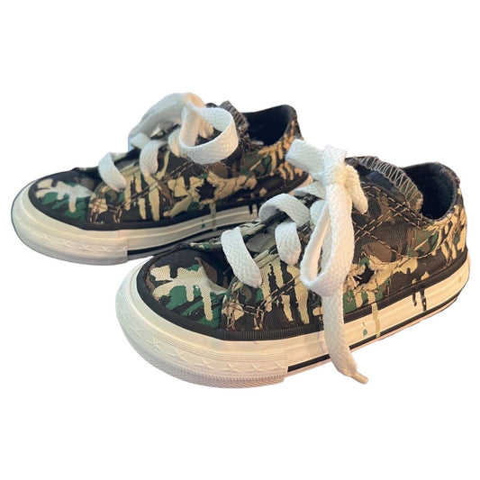 A pair of low-top canvas sneakers with a green, brown, and black camouflage pattern, featuring white laces and white rubber soles, shown against a plain white background.