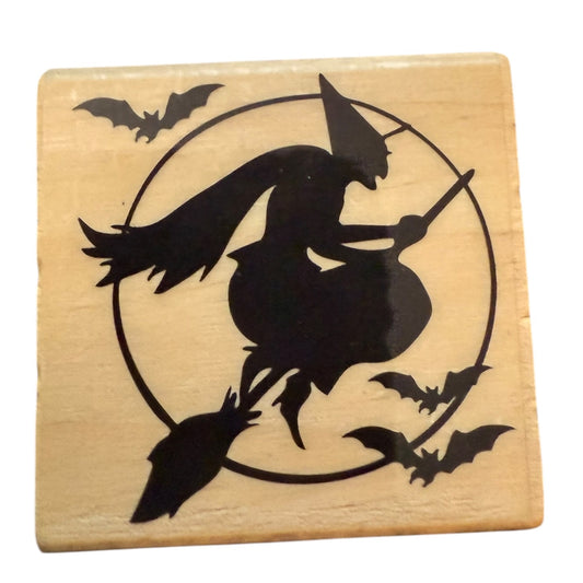 Wooden stamp with a black silhouette of a witch on a broom surrounded by bats on a beige background.