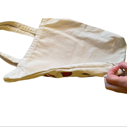 A cream-colored canvas tote bag with a hand holding the bottom edge to show its drawstring closure, isolated on a white background.