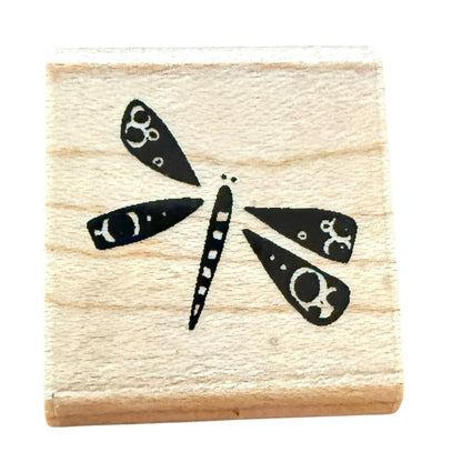 A square wooden rubber stamp block featuring a black dragonfly design with white circular details, isolated on a white background.