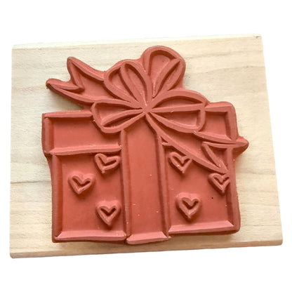 A rubber stamp gift box stamp with a large bow and small heart details, mounted on a light-colored wooden block.
