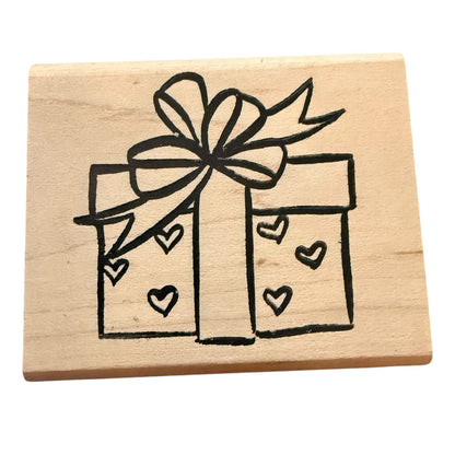 A square wooden stamp block featuring a black line-art design of a gift box with a bow and small hearts, shown on a plain white background.