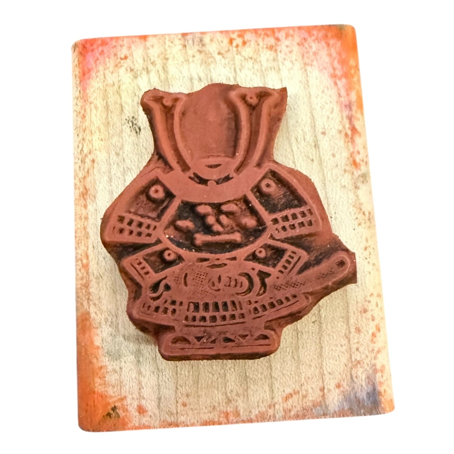 A terracotta-colored wooden stamp featuring an samurai warrior design, mounted on a rectangular, weathered beige wood block with orange-brown stained edges, set against a plain white background.
