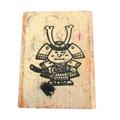 A square wooden stamp with a black ink illustration of a samurai character, shown on a plain white background.