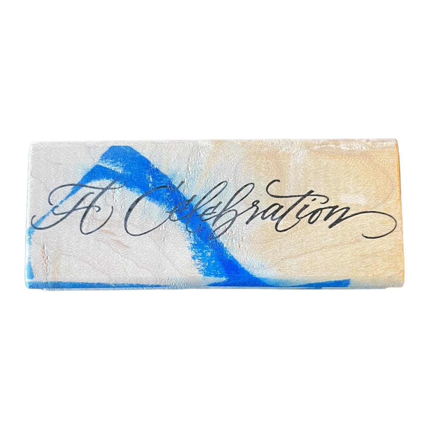 A rectangular wood stamp with the words 'A Celebration' written in black cursive script with a blue stain.