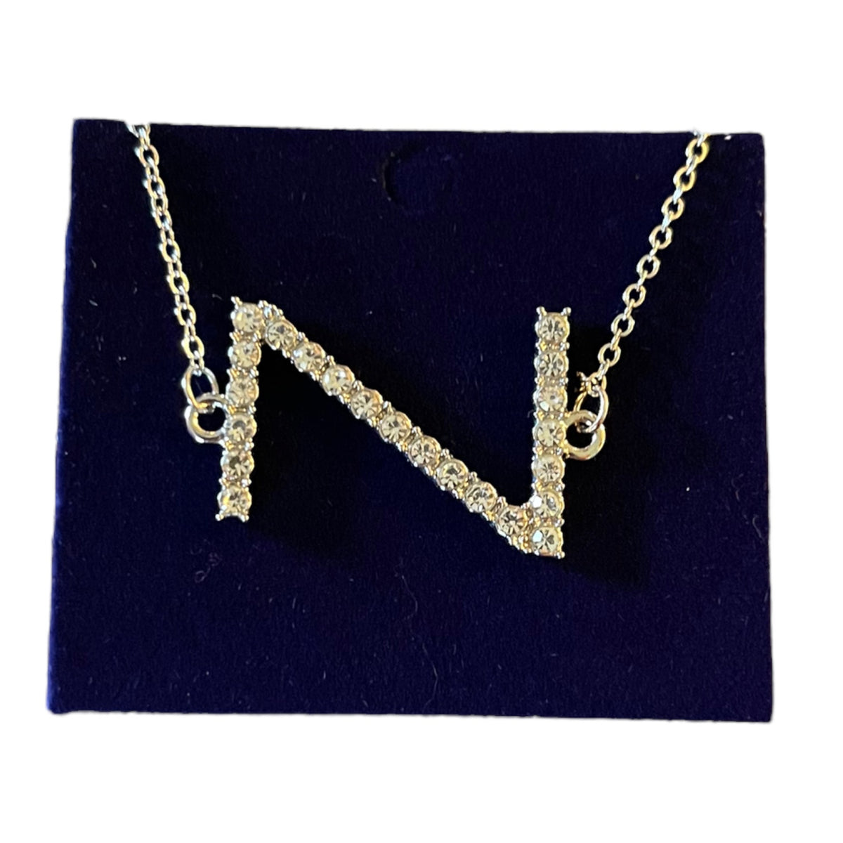 A silver initial necklace featuring the letter 'N' paved with clear stones, displayed on a dark navy blue velvet jewelry card against a white background.