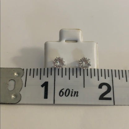 A pair of small round stud earrings with clear gemstones in silver-toned settings, displayed on a white stand above a metal ruler for scale.