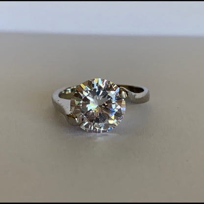 A round-cut clear solitaire ring set in a silver-colored metal band, resting on a neutral beige surface.