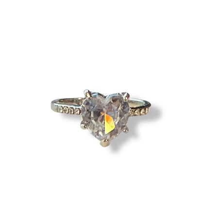 A heart-shaped clear gemstone ring with a silver-toned band featuring small accent stones on either side, shown on a plain white background with a soft shadow.