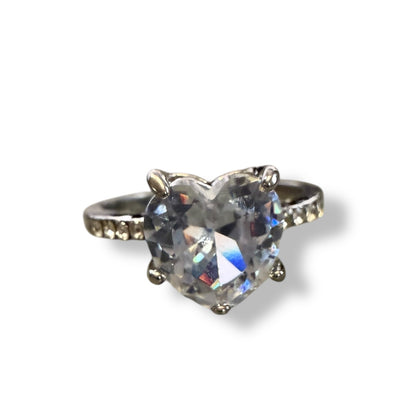A heart-shaped clear gemstone ring set in a silver-toned metal band with small accent stones on either side, shown on a plain white background.