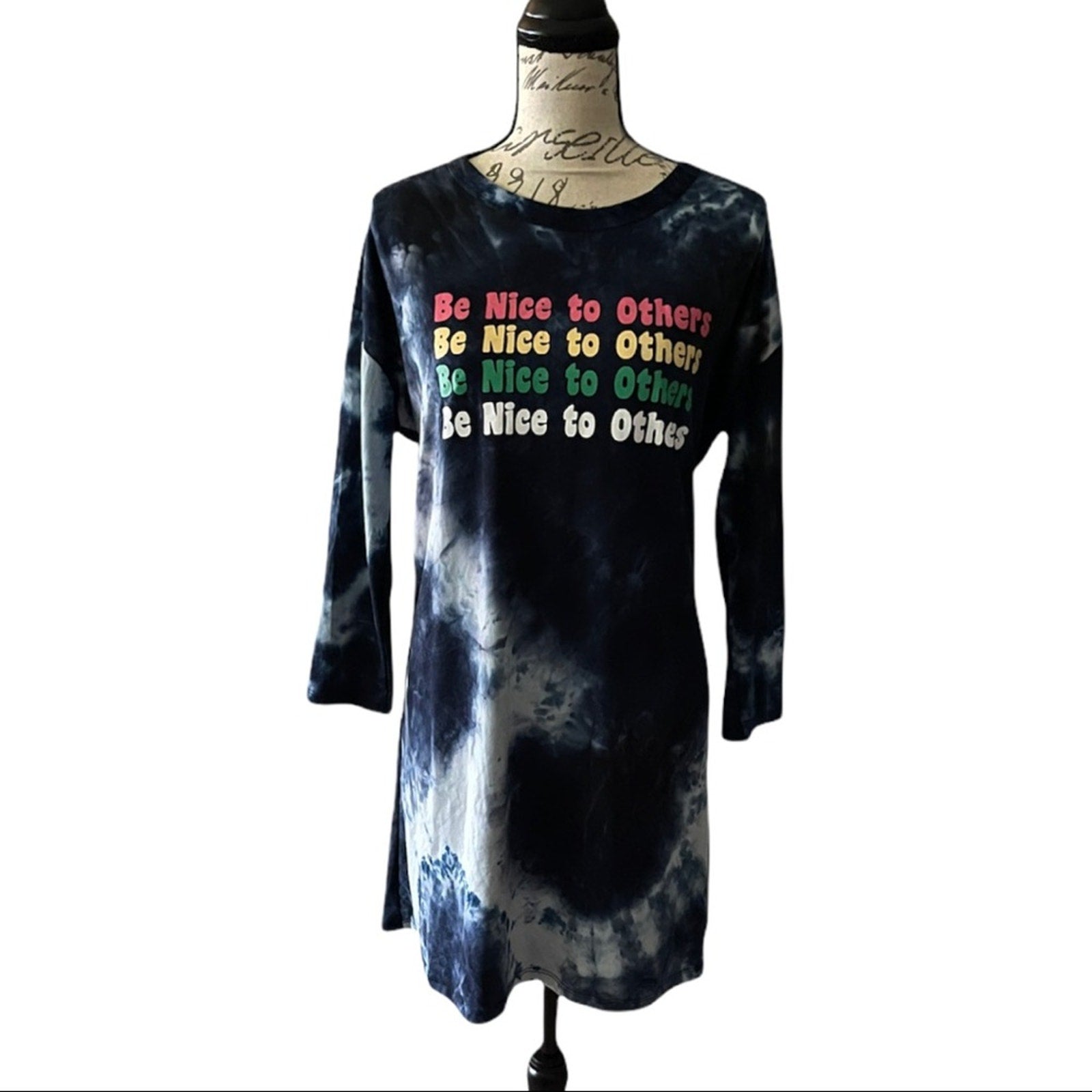 A long-sleeve tie-dye dress displayed on a dress form mannequin against a white background. The dress features a dark blue and light bue tie-dye pattern with a colorful text graphic on the chest that reads 'Be Nice to Others' repeated.