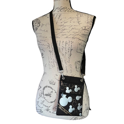 A black mini crossbody purse with Disney Mickey Mouse design displayed on a mannequin. The purse features  straps and multiple zippered pockets.